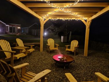A wooden pergola with string lights above wooden chairs around a fire pit in a garden at Gwennol Cottage Golden Grove near Llandeilo