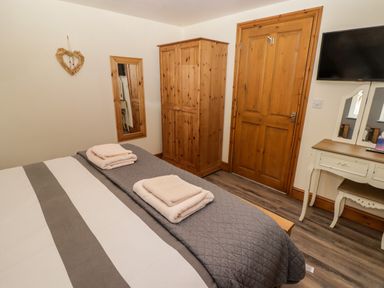 A bedroom with a bed with folded towels a wooden wardrobe a wall mirror a door a wall mounted TV and a dressing table with a mirror at Gwennol Cottage in Golden Grove near Llandeilo