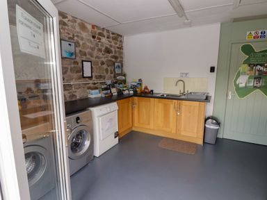 A laundry room with a washing machine, dryer, wooden cabinets, a sink, informational brochures, and a waste bin at Gwennol Cottage in Golden Grove near Llandeilo