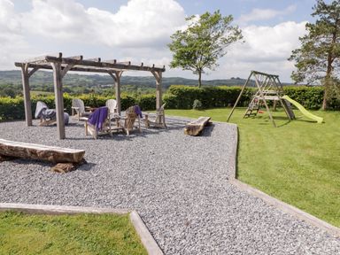 An outdoor seating area with wooden chairs under a pergola and a children's playset with swings and a slide on grass at Gwennol Cottage in Golden Grove near Llandeilo