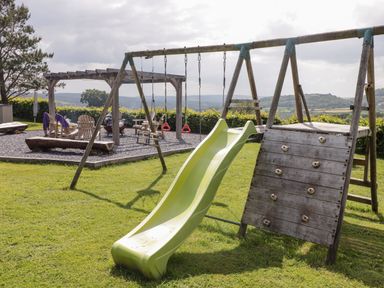 A playground with a green slide wooden climbing wall swings and a seating area with chairs under a pergola on grass at Gwennol Cottage in Golden Grove near Llandeilo