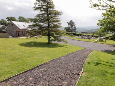 A garden with a gravel path leading to a lawn and a stone house with skylights and a playground with swings and a slide at Gwennol Cottage Golden Grove near Llandeilo