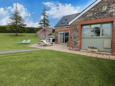 A stone cottage with a patio table and umbrella on a lawn with two lounge chairs and trees at Gwennol Cottage in Golden Grove near Llandeilo