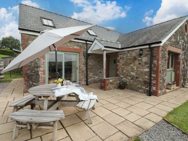 A stone cottage with patio doors wooden picnic table with umbrella and flowers on a paved patio at Gwennol Cottage in Golden Grove near Llandeilo