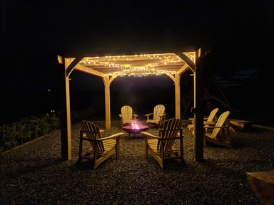 An outdoor seating area with wooden chairs under a pergola with string lights and a fire pit at Sewin Cottage Golden Grove near Llandeilo