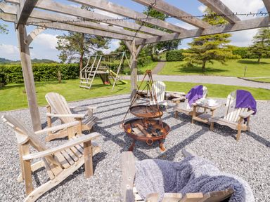 An outdoor seating area with wooden chairs around a fire pit under a wooden pergola with string lights and a children's playset in the background at Sewin Cottage in Golden Grove near Llandeilo