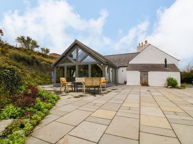 An outdoor patio area with wooden chairs and table next to a house with a glass extension and white walls at Balog Farm in Amlwch