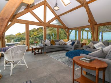 A sitting room with gray sofas pillows a white wicker chair wooden tables and large windows at Balog Farm in Amlwch