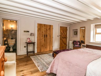 A bedroom with wooden floor beams wooden doors a bed with pink bedding a chair and a small table with a lamp at Balog Farm in Amlwch