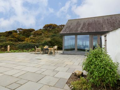 An outdoor patio with wooden chairs and a table next to a building with large windows at Balog Farm in Amlwch