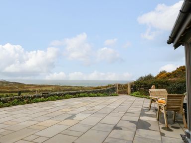 A patio with wooden benches overlooking a stone wall and grassy landscape with the sea in the distance at Balog Farm in Amlwch