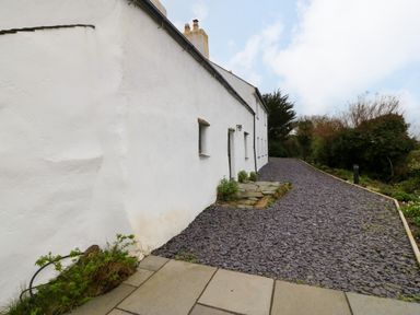 A white exterior wall of a building with small windows and a gravel pathway alongside trees at Balog Farm in Amlwch