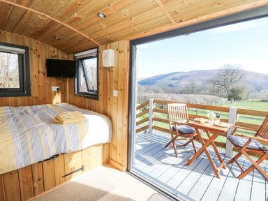 A small wooden bedroom with a bed towels and a wall-mounted television next to an open sliding door leading to an outdoor deck with a table and chairs at Elan in Rhayader