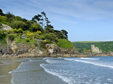 A beach with waves and rocky cliffs covered in trees and bushes with a stone ruin in the background at The Penthouse in Salcombe