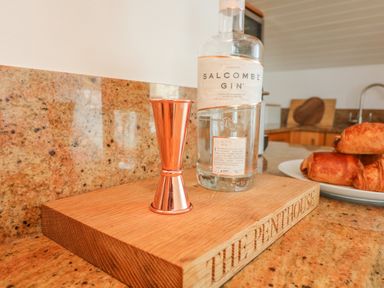 A kitchen counter with a bottle of Salcombe gin a copper jigger on a wooden block and pastries on a plate at The Penthouse in Salcombe