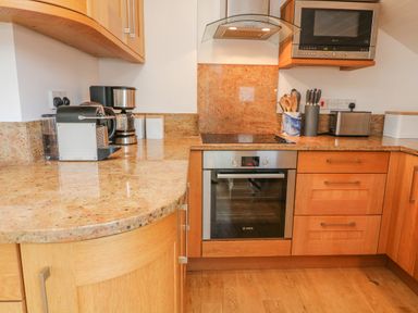 A kitchen with wooden cabinets, a granite countertop, a built-in oven, microwave, coffee makers, a toaster, and kitchen utensils at The Penthouse in Salcombe