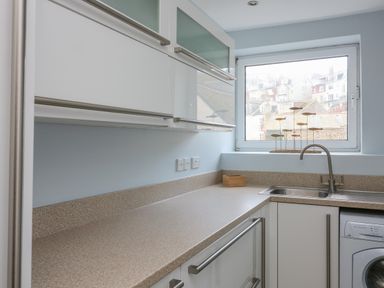 A kitchen with countertop sink washing machine cabinets and window showing buildings outside at The Penthouse in Salcombe