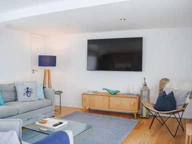 A living room with a wall-mounted TV a wooden cabinet a grey sofa with cushions a glass coffee table a blue floor lamp and a wicker chair at The Penthouse in Salcombe