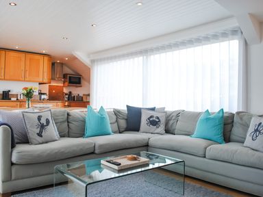 A living room with a gray sectional sofa with blue and patterned cushions and a glass coffee table with a tray and remote in front of sheer curtains and a kitchen with wooden cabinets at The Penthouse in Salcombe