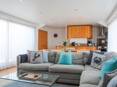 A living room with a grey sectional sofa with sea creature pillows and a glass coffee table in front of a kitchen with wooden cabinets and a dining table at The Penthouse in Salcombe