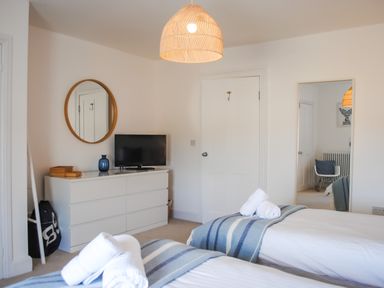 A bedroom with two beds with striped blankets and rolled towels a white dresser with a television and a round mirror on the wall at The Penthouse in Salcombe