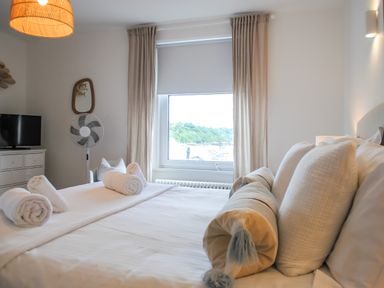 A bedroom with a bed and rolled towels near a window with curtains and a dresser with a fan and TV at The Penthouse in Salcombe
