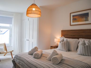 A bedroom with a wooden bed, striped bedding, rolled towels on the bed, a nightstand with a lamp, an armchair, and a window with white curtains at The Penthouse in Salcombe