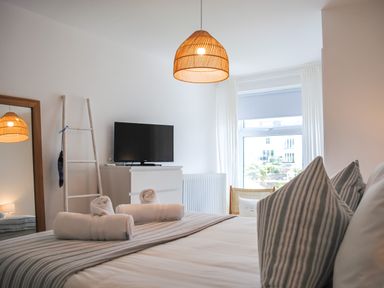 A bedroom with a bed with striped pillows and rolled towels a wooden ladder a television on a white dresser and a window with white curtains at The Penthouse in Salcombe