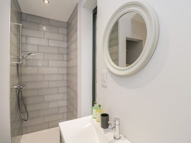 A bathroom with a shower area tiled with gray rectangular tiles and a white sink below a round white framed mirror at Lugg View in Cross Keys near Hereford