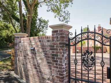 A brick wall with white caps and a black wrought iron gate in front of a gravel driveway and brick house at Lugg View in Cross Keys near Hereford