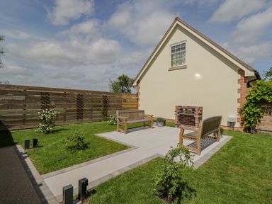 A garden with two wooden benches on a paved area and a small brick barbecue near a cream house at Lugg View in Cross Keys near Hereford