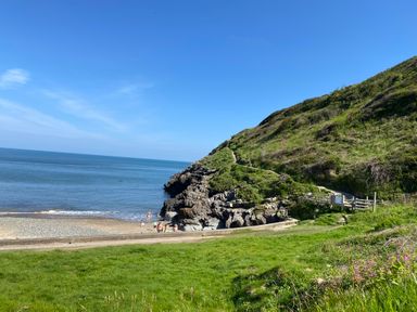 A coastal scene with a grassy hill, a rocky beach, and a blue sea at Dolwylan Barn in Cwmtydu near New Quay