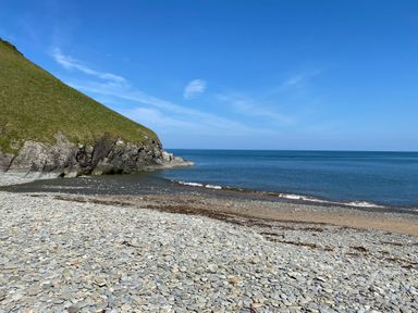A rocky beach with a grassy cliff and the sea under a blue sky at Dolwylan Barn in Cwmtydu near New Quay