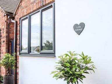 A white wall with a black heart-shaped sign saying Little Oakleigh next to a window and plants in pots at Oakleigh in Four Crosses