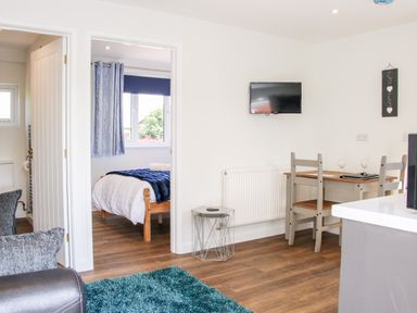 A living area with a small dining table and chairs a wall-mounted television and a bedroom visible through an open door at Oakleigh in Four Crosses