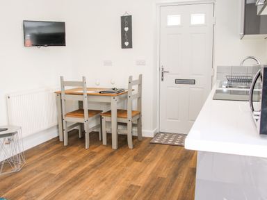 A dining area with a wooden table and four chairs a wall-mounted TV a white door and a kitchen counter at Oakleigh in Four Crosses