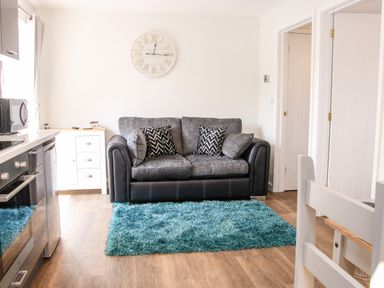 A living room with a sofa and two patterned cushions a blue rug a white clock on the wall and kitchen appliances at Oakleigh in Four Crosses