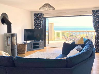 A living room with a blue sectional sofa a flat screen tv on a stand a wood stove and an open door leading to a balcony with chairs and a sea view at Ty Ni in New Quay