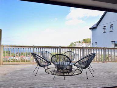 A wooden deck with five black wire chairs around a small round table and a railing overlooking houses and trees at Ty Ni in New Quay