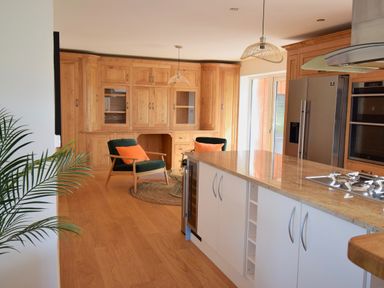 A kitchen with wooden cabinets an island countertop two chairs with orange cushions and a plant at Ty Ni in New Quay