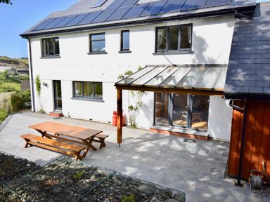 An outdoor patio with a wooden picnic table and benches beside a white house with solar panels on the roof at Ty Ni in New Quay