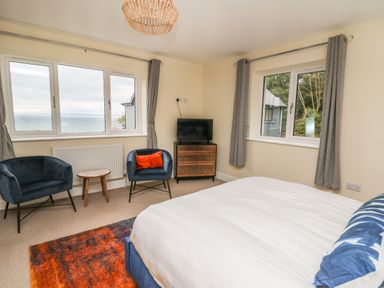A bedroom with a bed two chairs a small table a chest of drawers with a TV and two windows with curtains at Ty Ni in New Quay