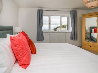 A bedroom with a bed with red pillows a window with blue curtains a lamp and a wooden wardrobe with a mirror at Ty Ni in New Quay