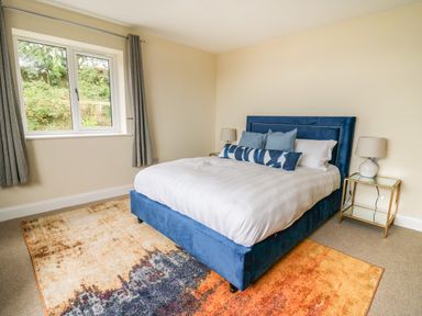 A bedroom with a blue upholstered bed a multicolored rug two bedside tables with lamps and a window with grey curtains at Ty Ni in New Quay