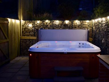 An outdoor hot tub with string lights on a stone wall and wooden fence at Ty Ni in New Quay