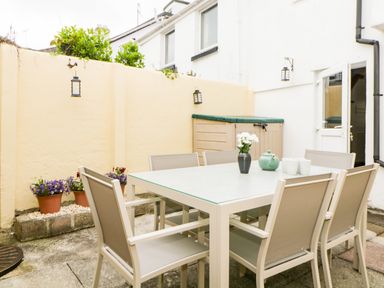 An outdoor patio area with a rectangular table and six chairs a vase with flowers and a teapot on the table potted plants along a wall and a door leading inside at The Yellow House 13 King Street Dawlish