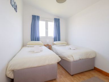A bedroom with two single beds with white bedding blue curtains and a small table with a lamp between them at 224 Atlantic Bays in St Merryn