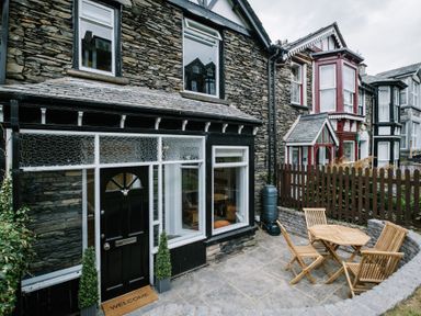 A stone house exterior with a black door and windows and a small outdoor seating area with wooden table and chairs at Craig Cottage in Bowness-On-Windermere