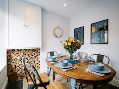 A dining area with a wooden table set with blue dishes and wine glasses with a flower vase in the center and wood logs stacked under a white cabinet at Craig Cottage in Bowness-On-Windermere