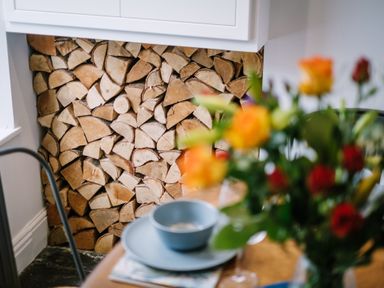 Stacked firewood in a white fireplace nook with a table holding a bowl and flowers in the foreground at Craig Cottage in Bowness-On-Windermere